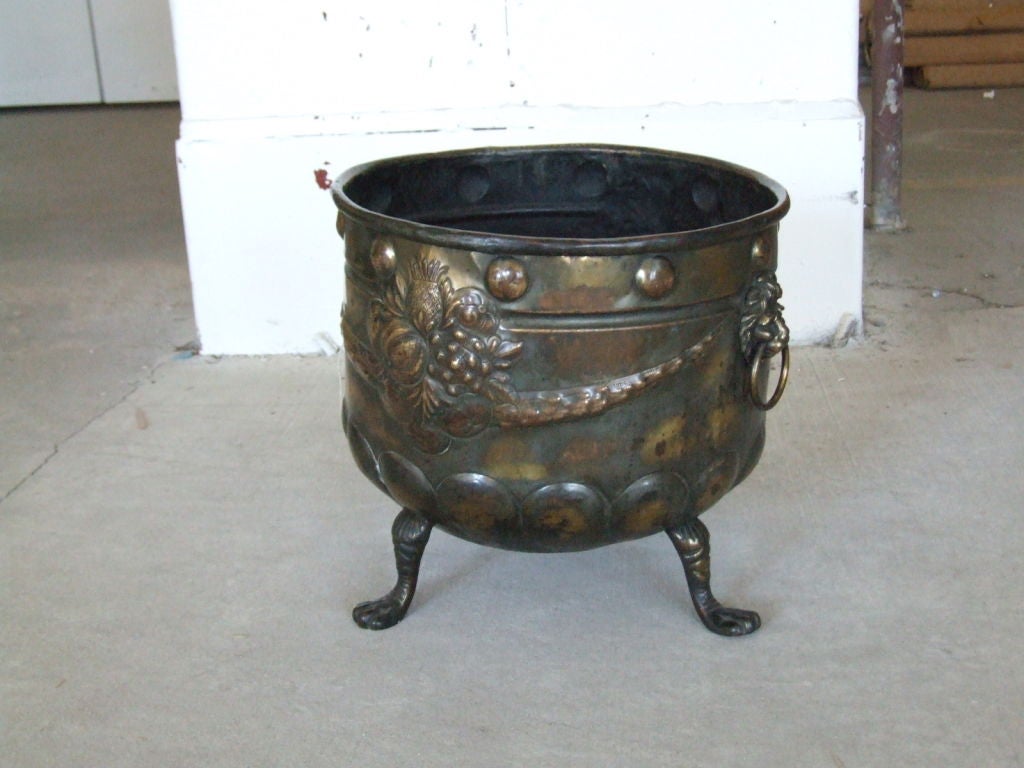 A 19th century brass Continental planter/kindling holder with rolled rim, lion head brass handles, repousee studwork and garlanded fruit decoration on three legs, the whole with bronze like patina.

firewood, kindling, log holder