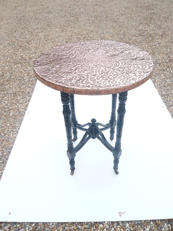 An English Aesthetic Movement ebonized table on turned legs joined by arched stretchers with finial, sumounted by a round, hammered copper top