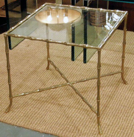 A chic 'Syrie' occasional table with an inset glass top and a square brass frame in a faux bamboo motif with an X-stretcher and petite flared feet. Manner of Bagues. By John Gerald, Inc.. U.S.A., circa 1950.