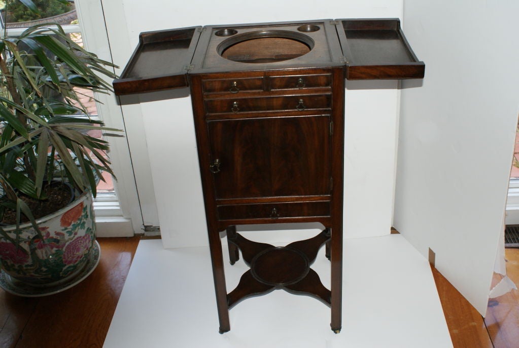 Of the period, a George III Flame Mahogany Gentleman's Wash Stand. The top opens from the center to show an opening that at one time held the wash bowl and shaving items. The bottom cross bar shelf is designed to hold a pitcher for water, in fine