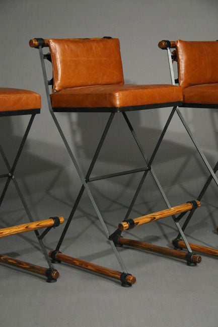 A pair of iron and wood frame bar stools with caramel leather cushions and wooden foot rests by Cleo Baldon for Terra.