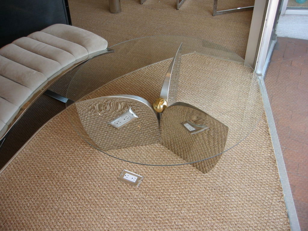 Round glass top cocktail table with a polished steel propellor shaped base. Brass ball in top center of base adding sculpture and dimension. A great functional table with a sense of humor.