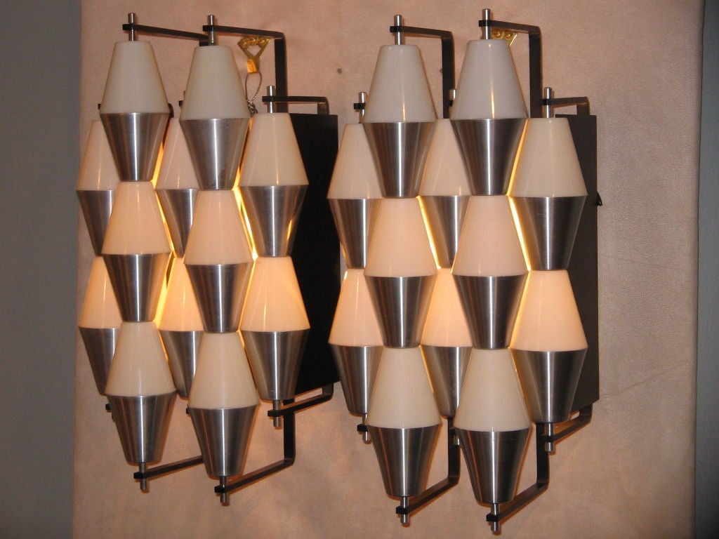 A pair of modernist wall sconces each featuring black metal frames which hold light diffusers which consist of rows of cone shaped cylinders in white plastic and aluminum. By RAAK, Holland, circa 1955. The sconces feature their original RAAK