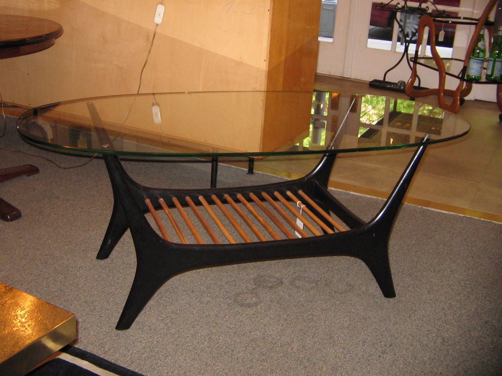 A cocktail table featuring a modernist shaped base with splayed legs in black lacquer with multiple cross stretchers in a blond wood. The table also features its original oval shaped glass top. By Alfred Hendrickx, Belgium circa 1955. Please see the