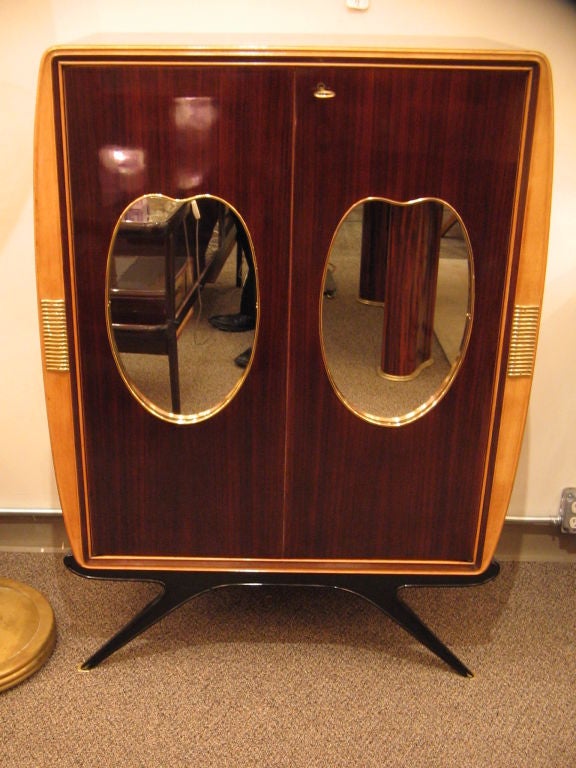 A two-door bar cabinet featuring a body in rosewood with waterfall edges, bombe shaped sides, a blond sycamore wood frame with applied brass detailing, splayed ebonized legs with brass feet and two inset brass framed front mirrors. The interior of