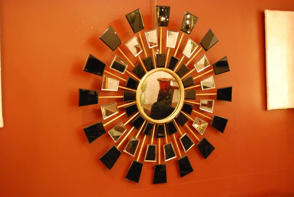 A large sunburst shaped wall mirror featuring a frame of gilded metal with a clear round central mirror and multiple arms holding rows of black or clear shaped pieces of mirror. Two mirrors available, sold individually<br />
France circa 1960.