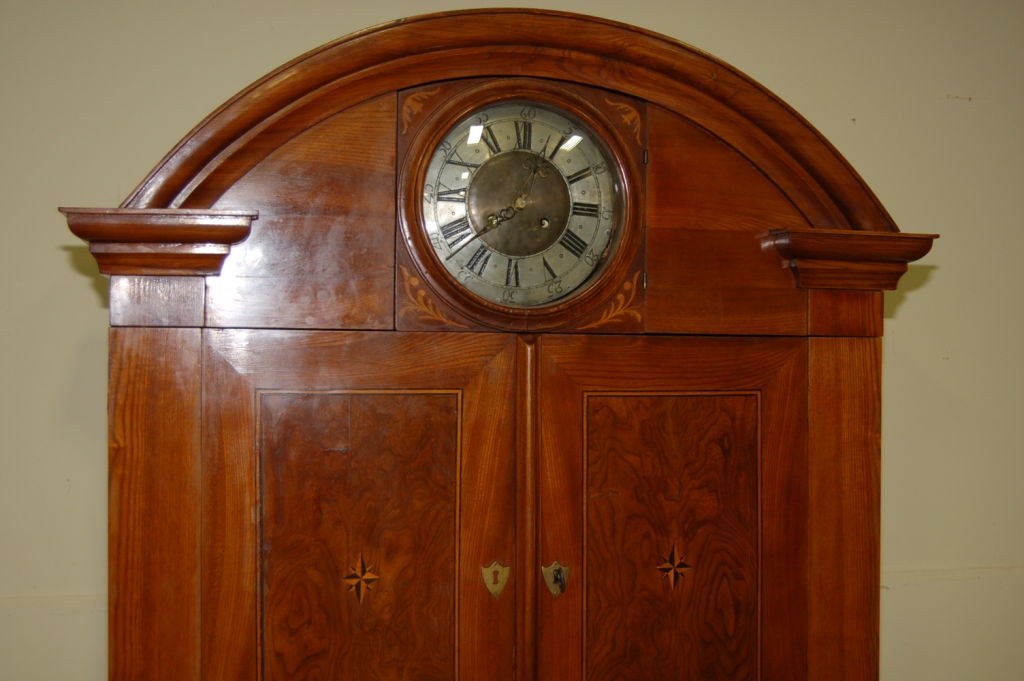 Early Biedermeier elm cabinet with 7-day clock, in working order solid and polished elm, circa 1825. Intarsias with elm, birch and moor oak. Orignal brass clockwork with 7 day movement and hour strike on a twin bell. Locks and hardware not original.