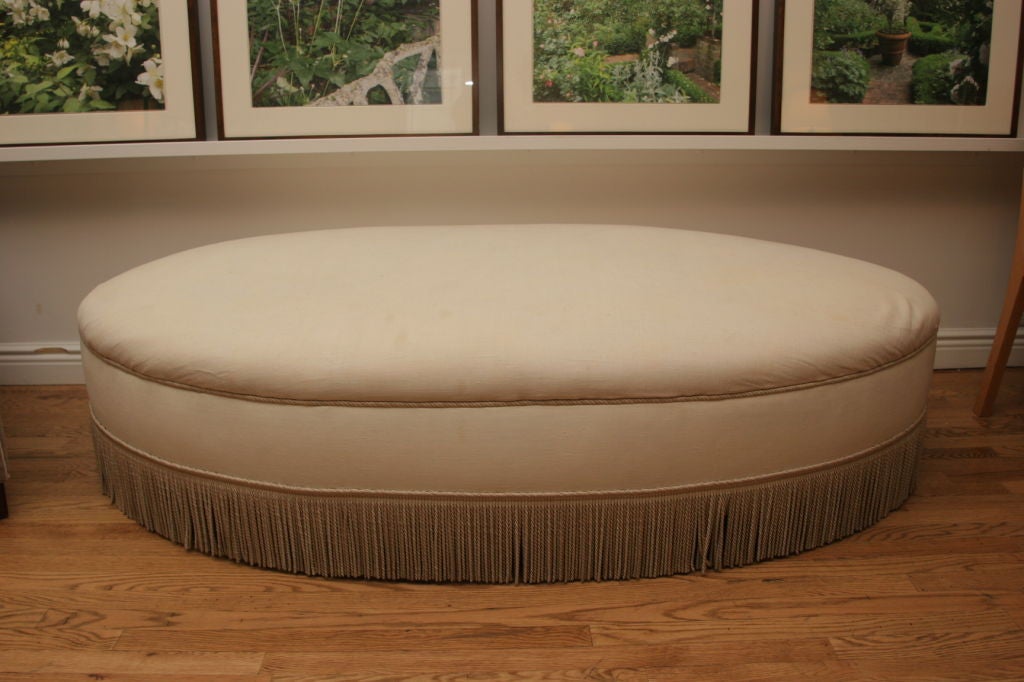 Large oval shaped ottoman upholstered in an antique linen with a silk bullion fringe.