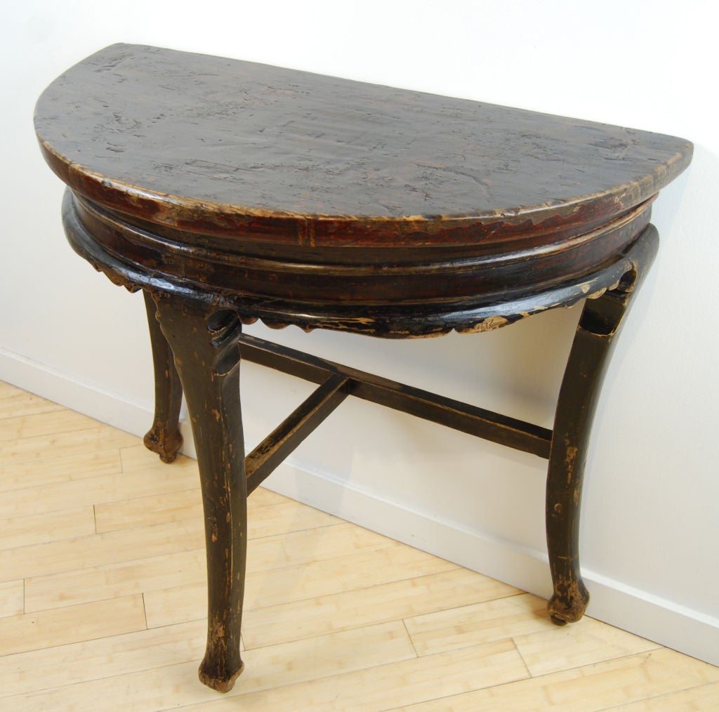 A Provincial Chinese elmwood half-moon table with traces of original red and black lacquer, an unusual old circular repair on top, and three legs ending in 