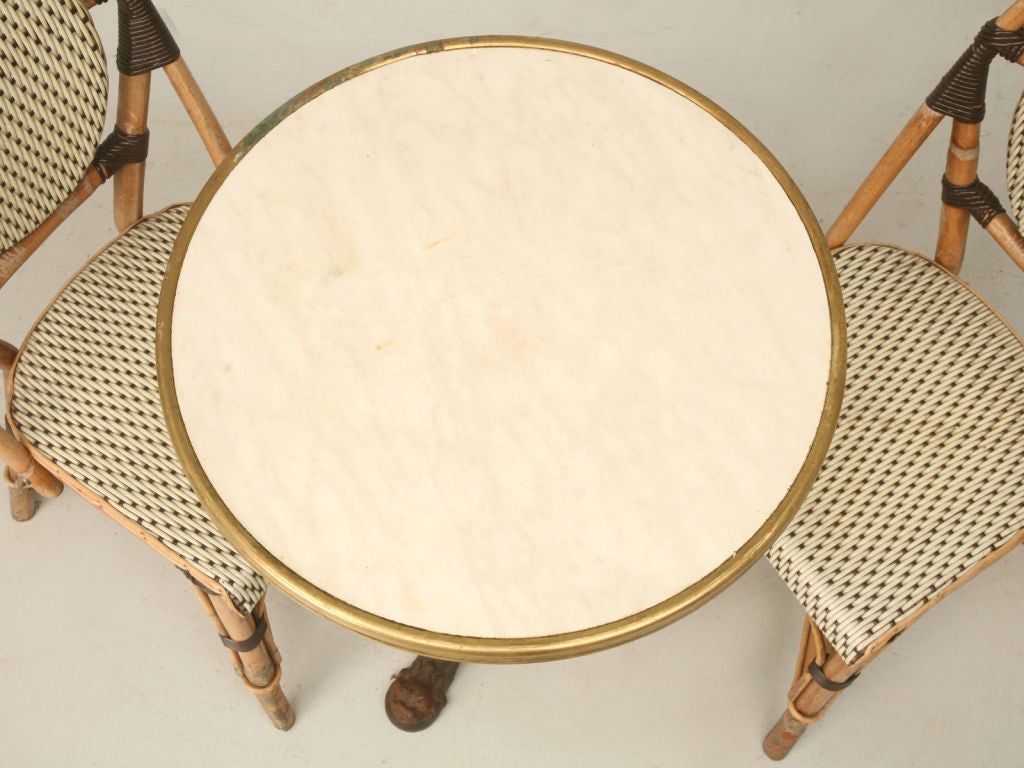 Vintage French iron bistro table that has a top with the look of marble but it is not stone with a brass trim. The chairs have bamboo frames with woven coated rattan backs and seats. The measurements below are for the table. The chairs are 36