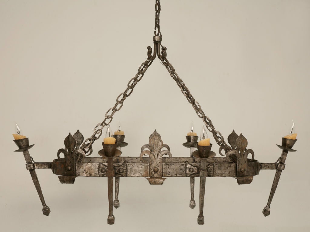 Spectacular vintage French hand-forged iron oval chandelier with six torch-form lights and fleur de lys details. Due to its mere size, it would be great over a kitchen island,   a pool table, in a wine cellar or ??? We have it pictured in a great