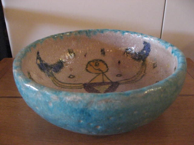 Whimsical Italian and mid-century bowl displays a lady in a dress holding two birds.  The design on the inside is a vibrant yellow, blue and purple while the exterior is turquoise and slightly textured.Signed with the donkey signature.