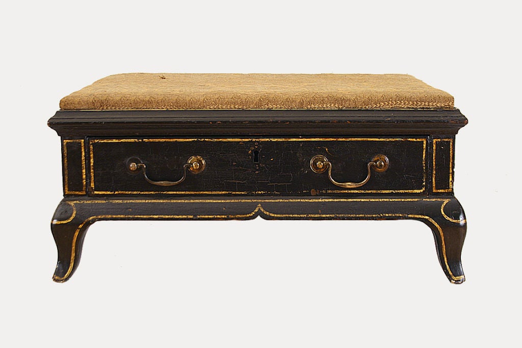 The upholstered top above a conforming case with one drawer, the body painted in black with gilt trim and ending in small pad feet, the bench used in a coach for storage and for resting the feet, retaining original brass hardware.
