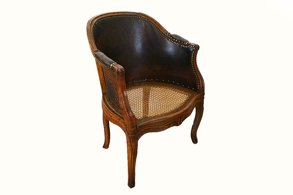 Of pegged construction and of barrel form, having a caned seat and back, the interior back upholstered in original, once red, leather and with a loose leather cushion, the channeled curved crest rail continuing past leather upholstered elbow pads to
