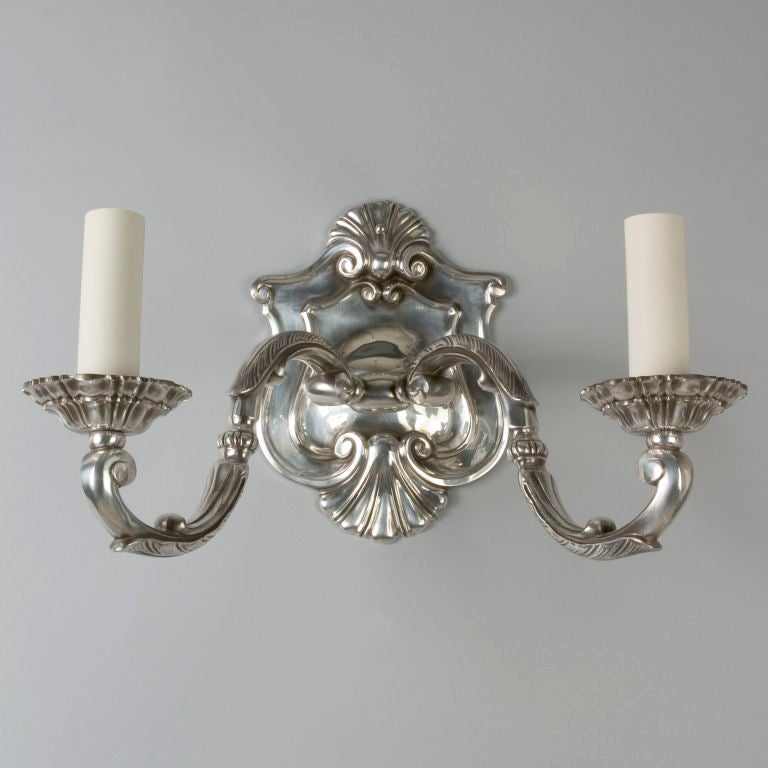AIS1838
A pair of large double light sconces in a silverplate over bronze finish. The shield-form backplates are finished above and below with shell motifs. The S-curving arms have scrolled terminals and foliate details. The bobeches are castings