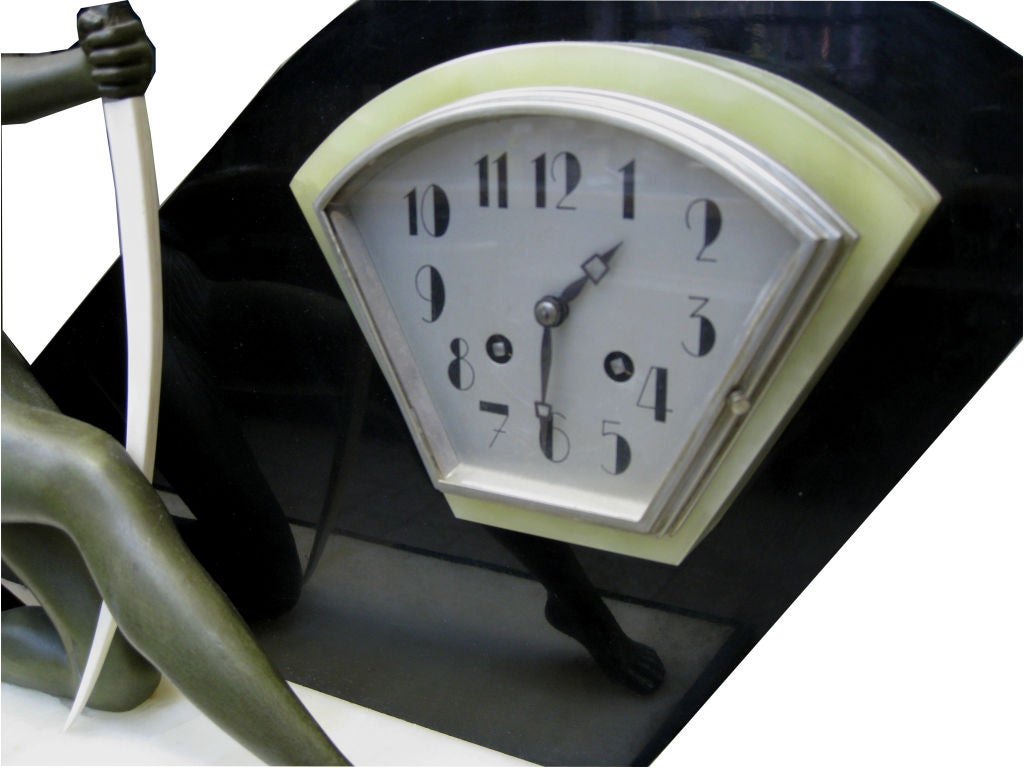 This large 1920’s French art deco clock features a bronze patinaed metal sculpture of Diana the Huntress with her ivory bow.  Diana kneels on the white marble top of a black marble base with sloping ends and inlays of translucent green and brown