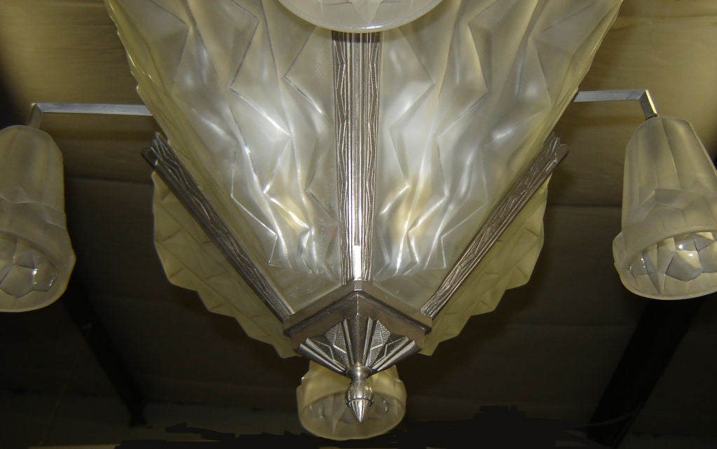 This 1930’s French art deco chandelier is by Degue and designed by Edouard Cazaux (1889 – 1974).  The nickeled metal body supports four large frosted glass “shields” and four curved arms, each hold frosted glass shades.  The geometrically formed