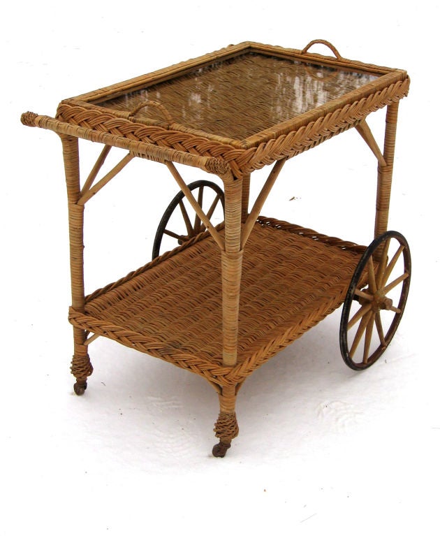 Bar Harbor wicker teacart in natural stained finish. Solid woven top and bottom shelves with wide braided borders. Large front wheels with wooden spokes, back feet with pineapple twist wrappings atop wooden casters. Removable wicker framed glass