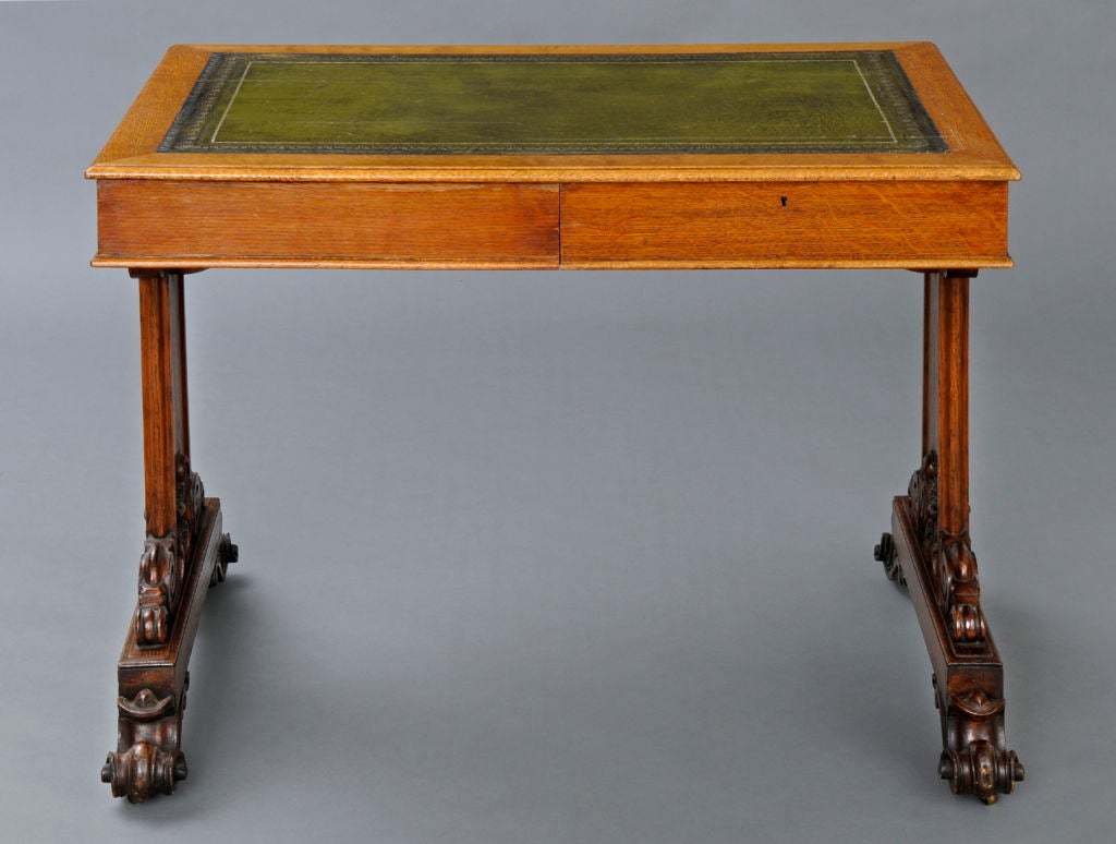 Early Victorian oak library or writing table with boldly carved end supports resting on carved scroll feet with recessed casters. There is one drawer on each side of the table. The writing surface is inset with green gilt-tooled leather.