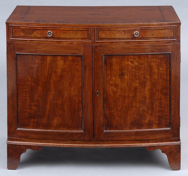 Georgian mahogany bow-fronted side cabinet, the top with wide mahogany cross-banding, two cock beaded drawers with ebony inlay and gilded knobs over two paneled doors enclosing an interior fitted with two compartments and three adjustable shelves,