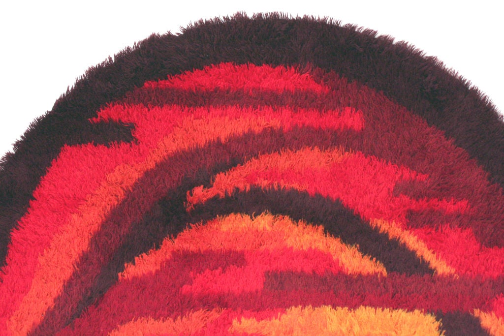 A large round Rya area rug in orage, red, and rust color palate and abstract cloudscape theme. Image is elliptical as it is shot on  a curved surface to feature entire graphic. Rug is 96