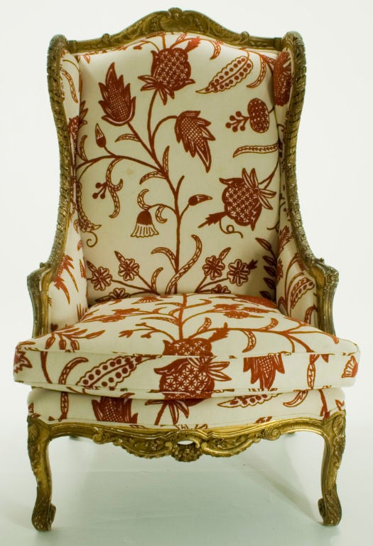 Outsized Alice in Wonderland-like Louis XV wing chair framed in ornately carved, kiln-dried, gold-leafed gesso  poplar. Upholstered in cream cotton canvas with a pattern of  large-print leaves,  vines and flowers in crimson wool embroidery. Labeled