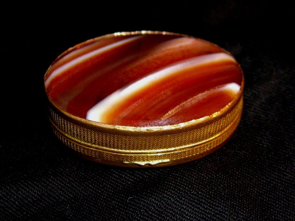 A beautiful pill or snuff box with beautiful agate top and bottoms. The top is a deep blre agate and the bottom is a more typical orange red agate. The metal is a gilt metal, possibly silver but not marked marked. The red agate side metal has some