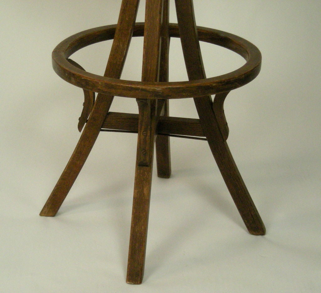 An adjustable height bentwood stool in oak, the swivel seat upholstered in brown leather with brass nail head decoration, over industrial cast iron hardware, on 3 legs joined by stretchers and circular foot rest.