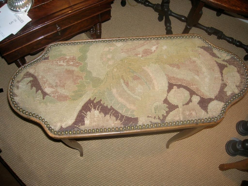 Elongated table with needlepoint top, edged with nailhead trim. Dusky rose, mauve, and sage yarns in needlepoint. Cabriole legs and semi-circular sides on top.