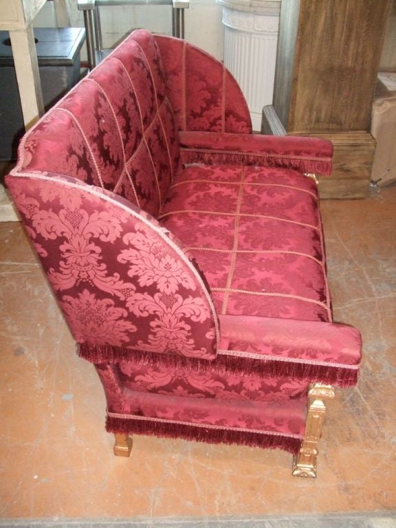 Upholstered settee in red damask with giltwood legs and fringe passementerie at base and arms. Matching gimp trimming on seat and back.