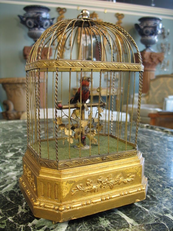 Singing mechanical bird in cage.  Giltwood base with neoclassical decoration, brass cage.  When activated, bird sings, its beak moves, its head turns and tail flaps.