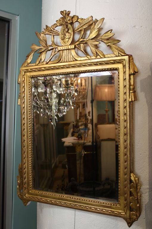 French Louis XVI period giltwood mirror with laurel leaf and floral crest, acanthus leave, tassels and other neoclassical details.  The glass has been replaced with a more recent bevelled mirror.
