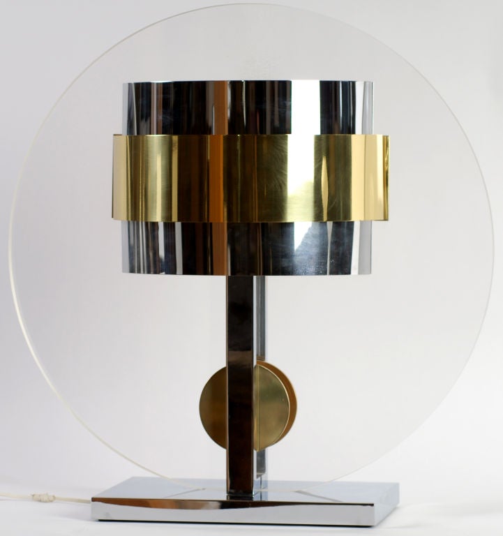 Upon a base and uprights of chromed steel, the thick Lucite disc of this lamp is mounted vertically.  On both sides, it has chrome and brass demi-lune shades, as well as suspended brass discs.