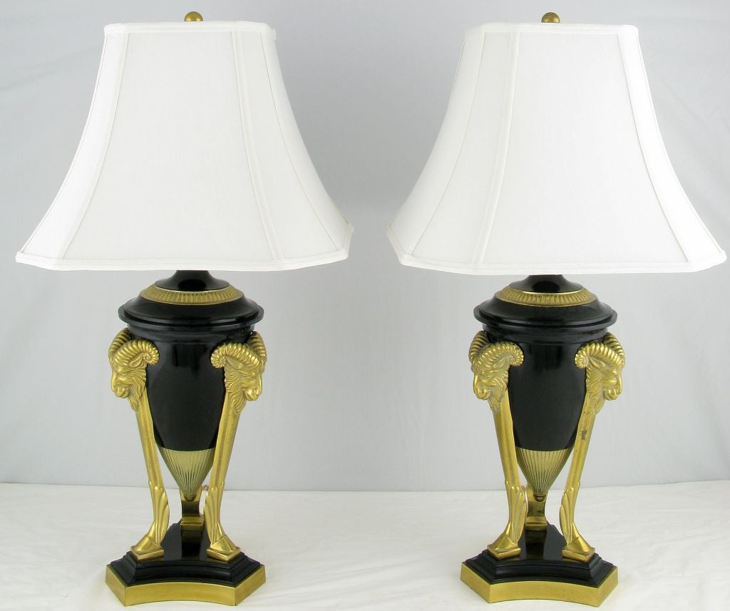 Elegant pair of lacquered metal and solid brass empire style table lamps. The conical center urn is supported by three hoofed pilasters, that are each crowned by a rams head. Sold sans shades.