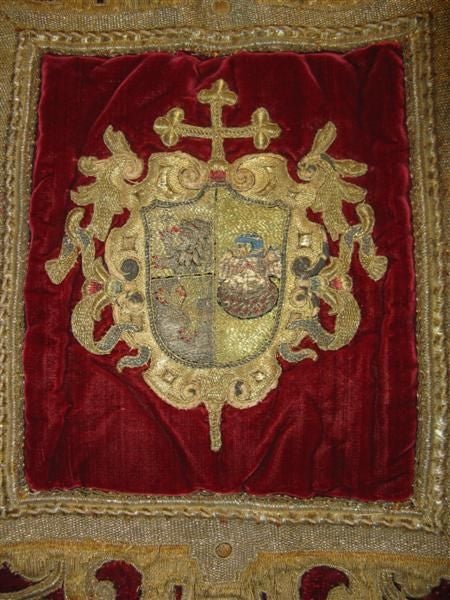 With it's crimson red background and gold metallic fringe, this wall hanging displays a family crest.  The crown at the top shows royal social ranking.  Metallic gold and silver needlework make up most of the embroidery.  The inner shield depicts a