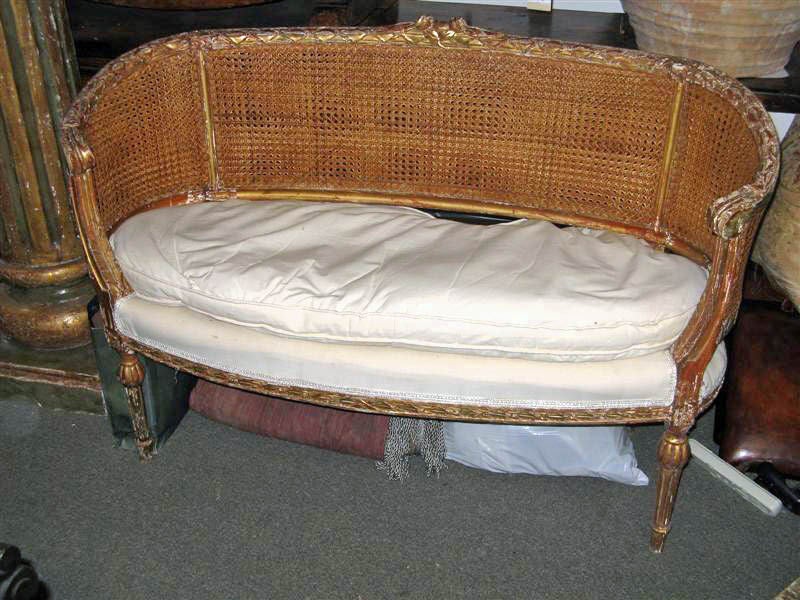 Charming giltwood 1920's French cane sofa/settee with upholstered cushion.  The wood carved decoration above the cane and along the apron has been painted and is worn with age. The fluted legs are adorned with a round shell like top.The double cane