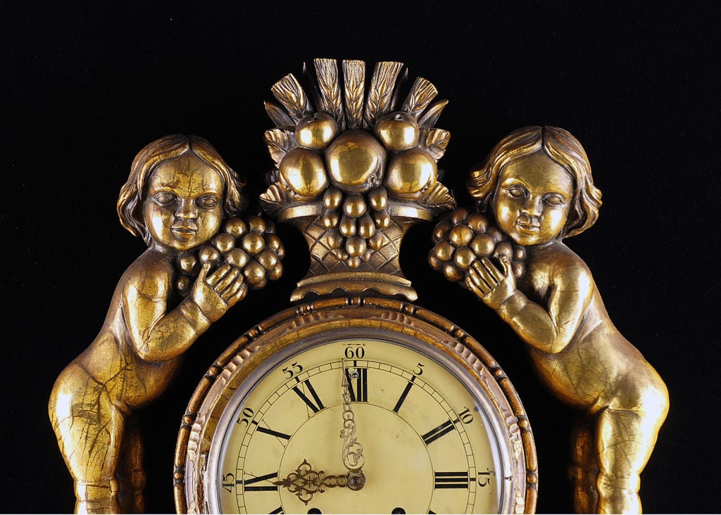 Intricately carved giltwood wall clock depicting the harvest.  Features clock face flanked by two children above carved and symmetrical flowing leaves and scrolls.<br />
<br />
For more great wall clocks, grandfather clocks and other decorative