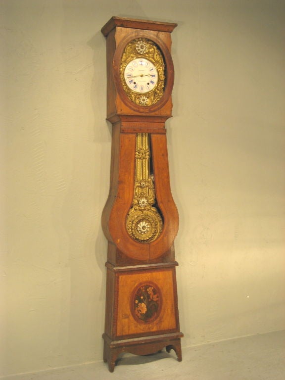 French comtoise with brass face surround and pendulum, beautifully embossed with peacocks, flowers, lyre, and rope chains. Gilt porcelain flowers punctuate the brass in varying scale. The case has painted line details and a bouquet-filled medallion.