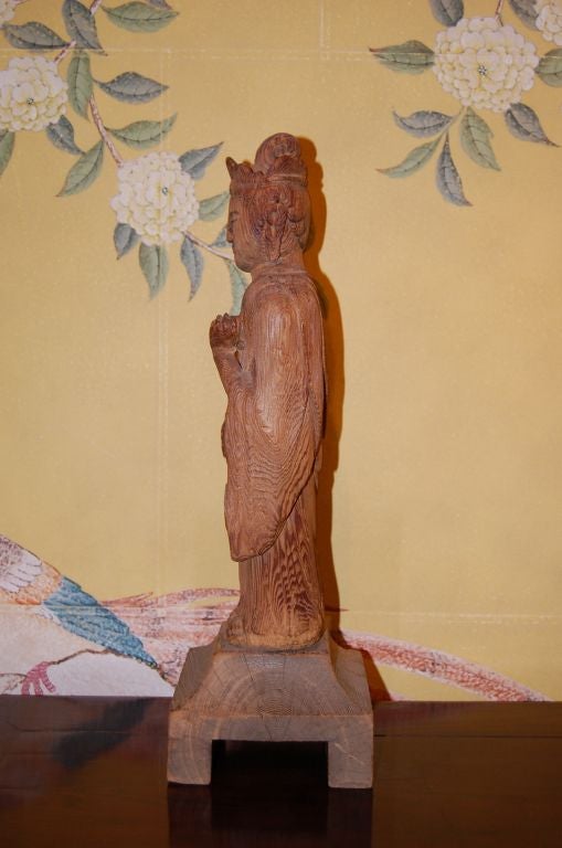 A simple wooden Japanese 18th century hand-carved statue of Kannon, the goddess of Mercy. The figure has a peg at the bottom, and this peg fits into the wooden base.
She is standing in a robe, with one hand down, and one up and facing the viewer.