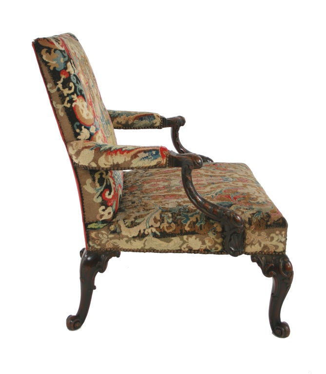 A George III carved mahogany Gainsborough chair with needlework upholstery, the downswept molded arms carved with C-scroll and cabochon clasps, the cabriole legs similarly ornamented with scrolled feet.  Needlework of the same period. Provenance: 