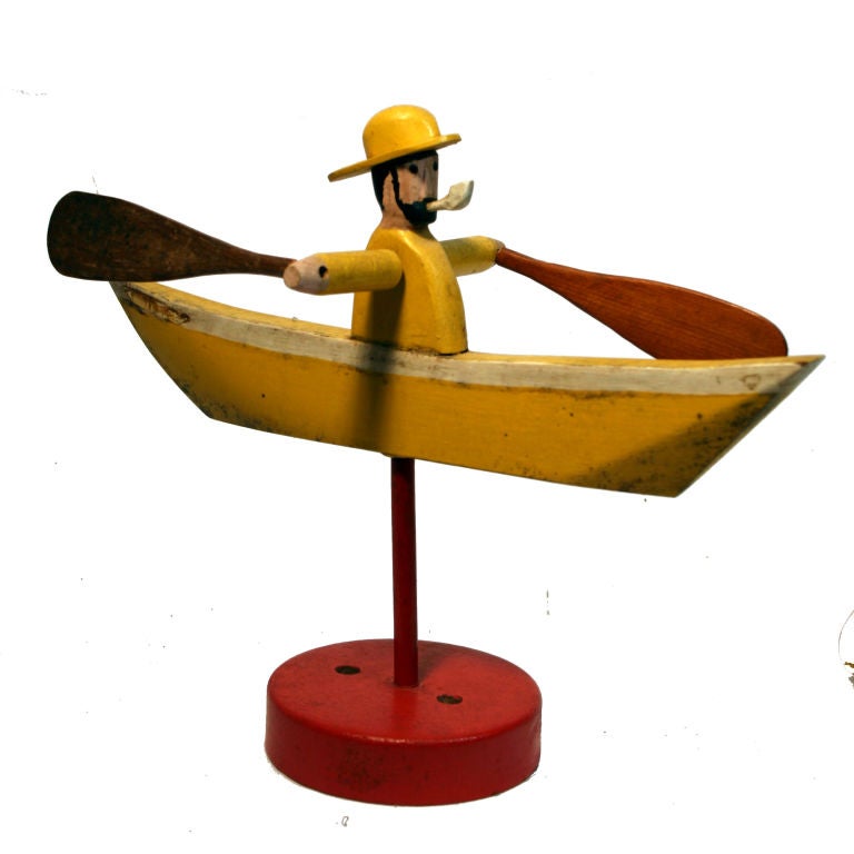 Made in Nantucket, this whirligig is made of carved wood that is painted yellow, white, and black with a red base, and depicts a sea captain in a boat. Whirligigs were creatively used to keep unwanted animals out of the garden. They were also called