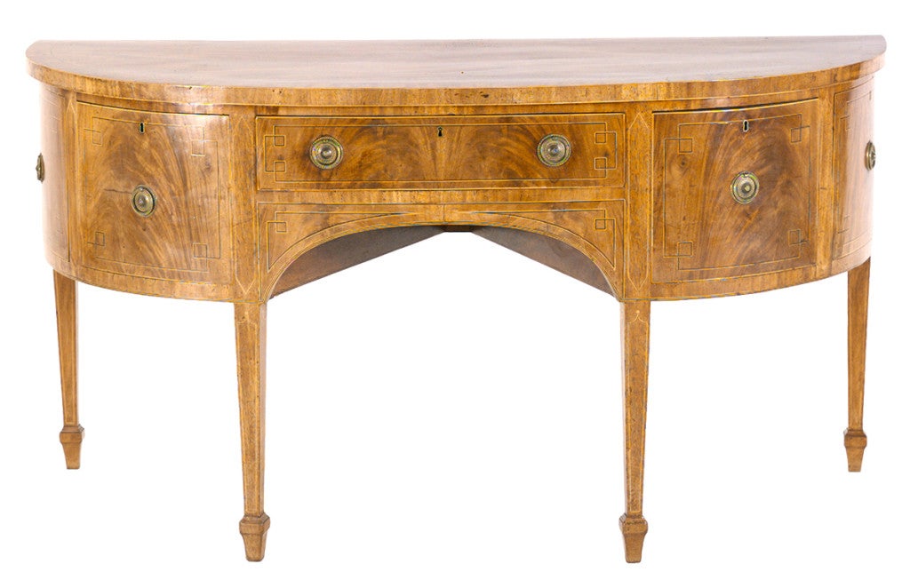 A Sheraton period mahogany sideboard with faded, blonde color. The d-shaped top is cross-banded and inlaid with stringing. The central drawer is flanked on each side by a drawer and cupboard door, all of which are inlaid with geometric stringing.