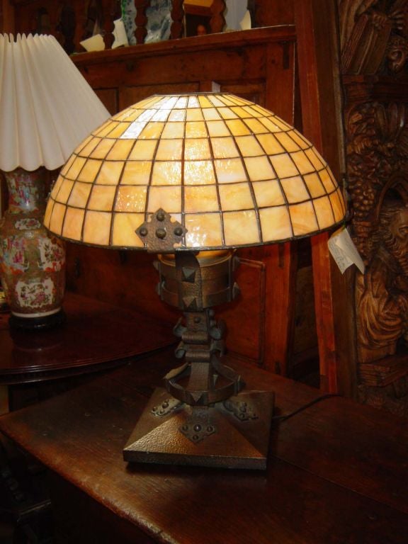 A large and impressive table lamp with copper and bronze base with hammered surface and strap decoration. with its original<br />
leaded caramel glass shade with great irridescence.  Originally<br />
made for keroscene, it is now electrified. 
