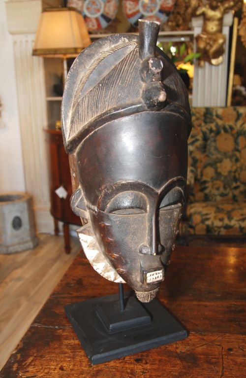 Tribal mask of the Baule People of the Ivory Coast in Africa. Sculptural rendition in hand carved and painted wood with a headdress and geometric flanges. There is ceremonial facial scarring depicted as well.