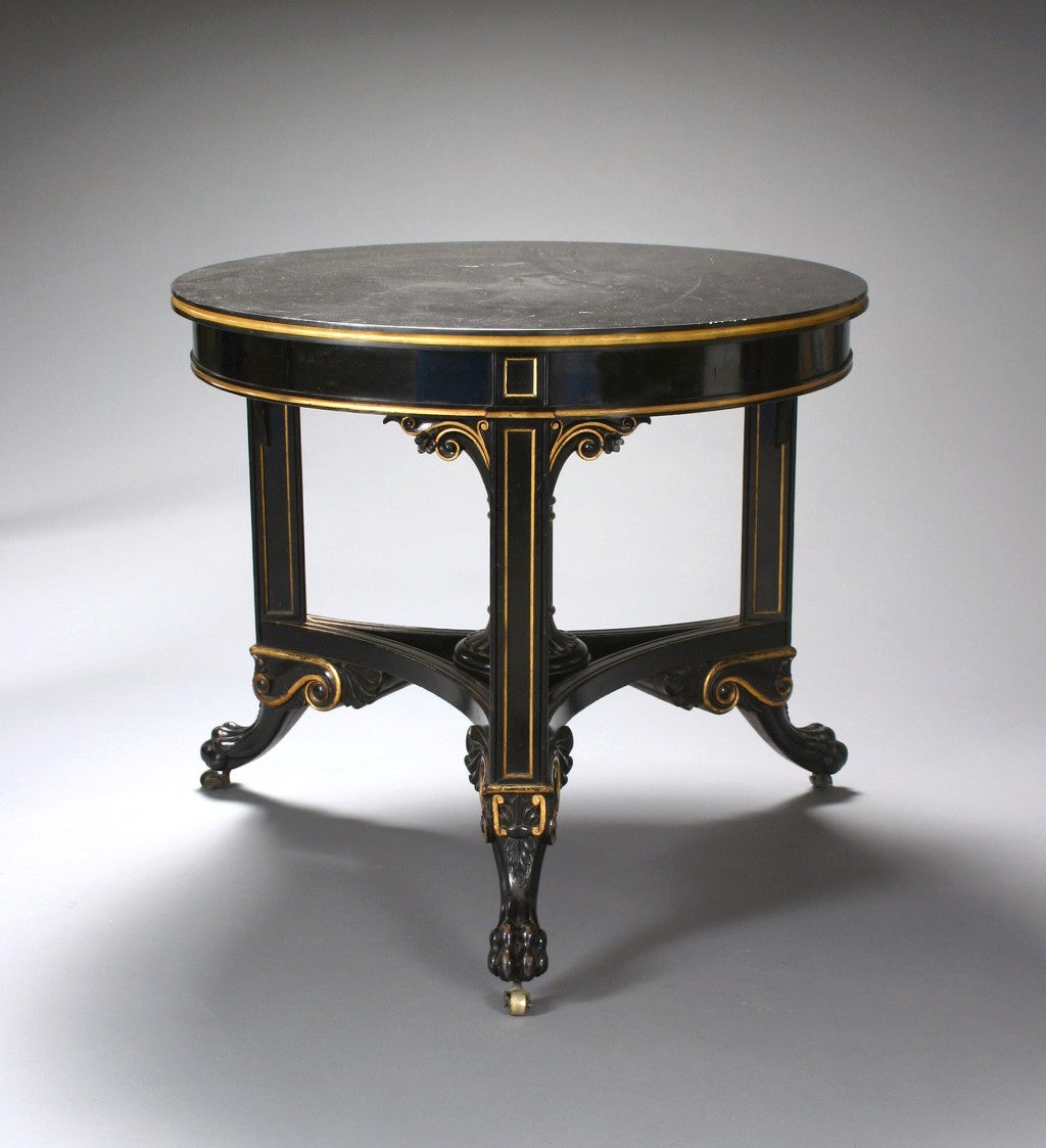 The circular black marble top with a grooved and gilded edge above the gilt edge apron, on three paneled triangular legs with flower-head brackets joined by a a concave sided triangular stretcher centering a column, ending in stylized winged lion