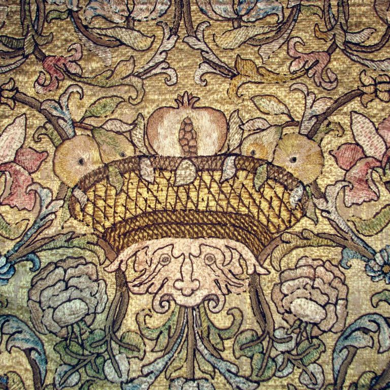 A delightful late 17th century Italian silk floss panel with scrolling vines and flower heads with three grotesque faces across with baskets filled with pomegranates on their heads.