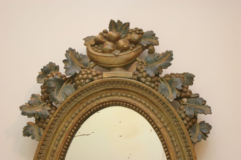 A Swedish Late Gustavian giltwood and painted candle mirror, Early 19th Century, probably from Gothenberg; the oval mirror plate surrounded by a carved molded frame and surmounted with an urn filled with fruits with flowing vine leaves and berries,