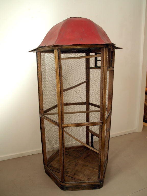 This free standing wood hexagon bird cage is wrapped in fine chicken wire with a top made of hammered tin and painted red. Each door opening is framed in the same wood as the birdcage together with a wood latch. The interior base is wood. There are