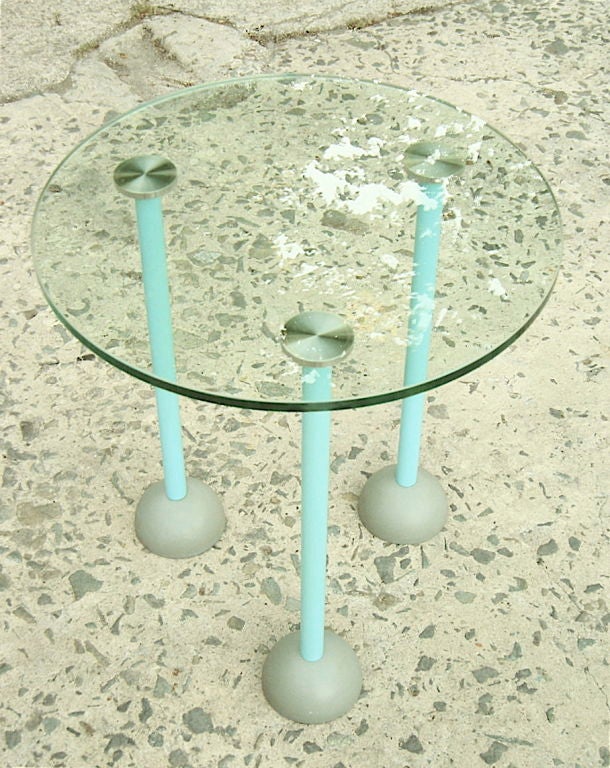 Glass top side table by Memphis, 1987. Date and Item number stamped on bottom.  Practical design with low maintenance glass surface.  Despite its handy dolly wheels it is very stable. 
Whimsical design references toilet plungers w/Aqua enameled