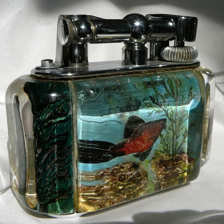 A silver-plated 'Aquarium' table lighter in 'Half-Giant' size, the perspex body enclosing decoration of fish, in pale blue-grey colour, and chrome plated lift-arm marked DUNHILL. <br />
<br />
<br />
<br />
Notes: The ‘Aquarium’ lighters were